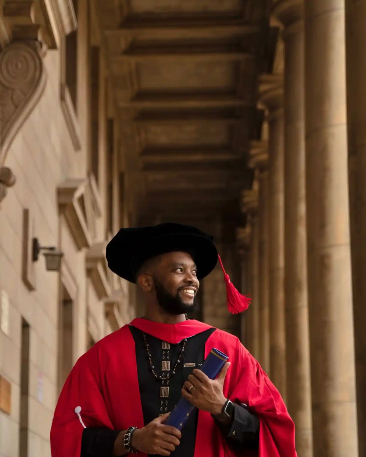Dr Kalonji smiling in red and black doctoral robes and a black bonnet, holding his degree scroll under a stone colonnade